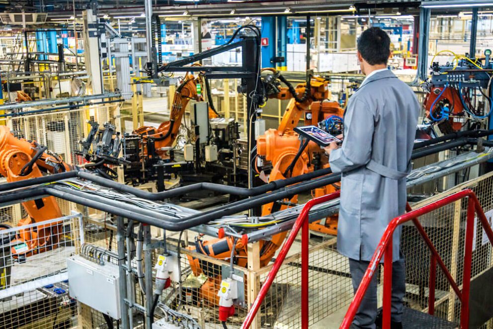A man is checking a clipboard while watching over an industrial warehouse, with machines and robots on an assembly line.